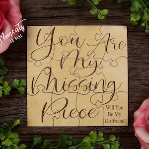 May include: A wooden puzzle with the words "You Are My Missing Piece" engraved in a cursive font. The puzzle also includes the text "Will You Be My Girlfriend?" in a smaller font. The puzzle is surrounded by greenery and pink flowers.