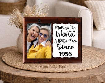 Può includere: Una targa rettangolare in legno scuro con una clip portafoto sul lato sinistro che trattiene un'immagine di due persone sorridenti. A destra, un pannello bianco smerlato mostra il testo nero: Making The World A Better Place Since 1956.