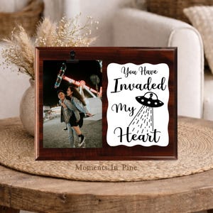 May include: A rectangular dark brown wooden photo frame with a black metal clip on the left side holding a photograph of two people at a fair. The right side features a white scalloped plaque with the black text You Have Invaded My Heart.
