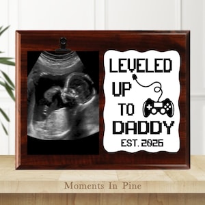 May include: A dark wood picture frame with a sonogram image and a white graphic that reads "LEVELED UP TO DADDY EST. 2026" with a controller icon. The frame is displayed on a light brown surface.