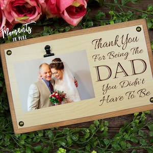 May include: A wooden picture frame with a photo of a couple, held by a black clip. The frame has the text "Thank You For Being The DAD You Didn't Have To Be" engraved on it. The frame is surrounded by pink roses and greenery.
