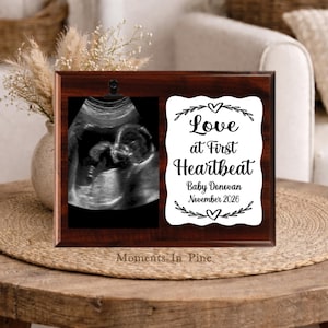 May include: A dark wood plaque featuring a black metal clip holding a black and white ultrasound image. Next to the image is a white scalloped sign with the text "Love at First Heartbeat", "Baby Donovan", and "November 2026" in black script.