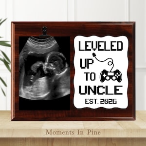 May include: A wooden picture frame with a sonogram image and the text "LEVELED UP TO UNCLE EST. 2026" in black. The frame has a dark brown finish and a clip to hold the sonogram.