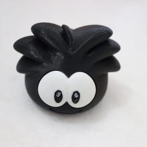 May include: A black, 3D-printed character with large white eyes and a small smile. The character has a spiky, rounded head. The object is on a white surface.