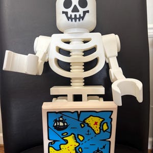 Puede incluir: Una figura de esqueleto grande, blanquecina, con una cara de calavera negra y una base de bloque de madera. El bloque presenta un mapa azul con masas terrestres amarillas, una calavera y un barco. La figura recuerda a un popular juguete de construcción.