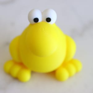 Può includere: Una statuetta a forma di rana gialla brillante, in stile cartone animato, con grandi occhi bianchi e pupille nere. La rana ha un corpo arrotondato, braccia piccole e piedi palmati. Il giocattolo è fatto di materiale liscio.