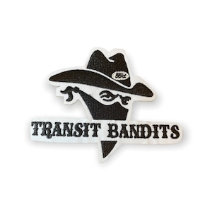 May include: A white patch with black embroidered text and a graphic. The graphic is a bandit wearing a hat and eye mask. The text reads "TRANSIT BANDITS" in a bold font. The patch has a clean, modern design.