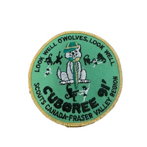 May include: A round green Cuboree patch with gold trim. The patch features a white cartoon wolf wearing a green hat and scarf. Text includes "Look Well O'Wolves" and "Scouts Canada-Fraser Valley Region".