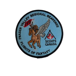 May include: A round, embroidered patch with a light blue background. The patch features a brown horse with wings, ridden by a figure, and a red flag. Text includes "Fraser Valley Regional Beaveree", "Flights of Fantasy", and "Scouts Canada".