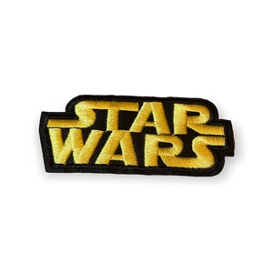 May include: Embroidered patch featuring the words "STAR WARS" in gold thread on a black background. The patch has a rectangular shape with rounded corners. The letters are bold and easily readable.