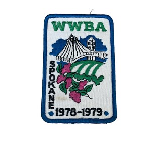 May include: Embroidered patch with a blue border and a white background. The patch features the text "WWBA" in green, the word "SPOKANE" in black, and the years "1978-1979". A stylized image of a building and a bridge is also present, along with pink flowers.