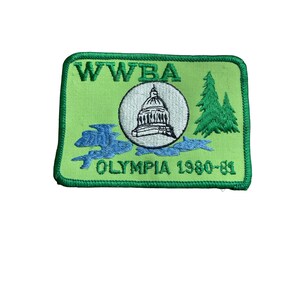 May include: A rectangular green patch with the text "WWBA" at the top. The patch features a white circle with a building illustration, blue water, and green trees. The text "OLYMPIA 1980-81" is at the bottom.