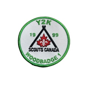 May include: Circular patch with a white background and a green border. The patch features the text "Y2K" above a black triangle with a red maple leaf and the text "SCOUTS CANADA". The text "WOODBADGE 1" is also present.
