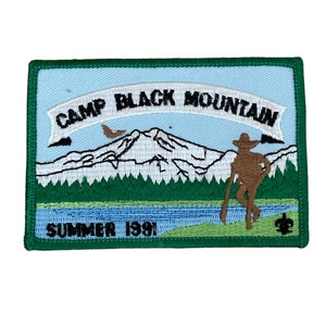 May include: Embroidered patch with a light blue sky background and a green border. The patch features a mountain scene with the text "CAMP BLACK MOUNTAIN" and "SUMMER 1991". A brown figure of a person is standing near a lake.