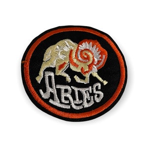 Op de afbeelding: Geborduurde ronde patch met een ramontwerp in oranje en crème op een zwarte achtergrond. De ram is omgeven door een oranje rand, met het woord "ARIES" in witte letters eronder. Ideaal voor het personaliseren van kleding of accessoires.