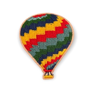 Puede incluir: Parche bordado con forma de globo aerostático. El globo presenta un diseño colorido con secciones rojas, amarillas, azules y verdes. El parche tiene un borde marrón claro y una pequeña cesta en la parte inferior.