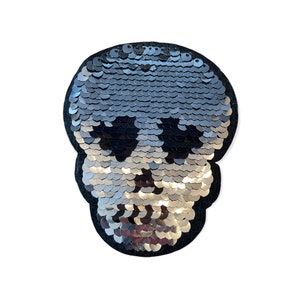 Puede incluir: Un parche con forma de calavera con lentejuelas plateadas, contorneado en fieltro negro. Las lentejuelas crean un efecto degradado, pasando de gris oscuro en la parte superior a plateado en la inferior. El parche es un artículo decorativo, adecuado para ropa o accesorios.
