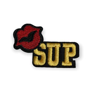 May include: Embroidered patch set featuring a red lip design and the word "SUP" in gold lettering on a black background. The patches are likely for clothing or accessories.