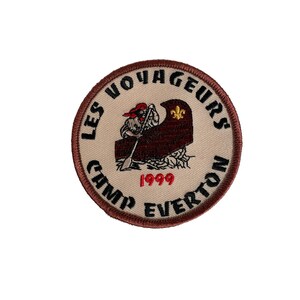 May include: Round embroidered patch with a brown border and a tan center. The patch features a canoe with a person paddling, the text "LES VOYAGEURS CAMP EVERTON 1999", and a gold fleur-de-lis.