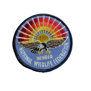 May include: Embroidered patch featuring a soaring eagle with outstretched wings, set against a rising sun in shades of yellow, orange, and red. The patch is circular, with the text "NATIONAL WILDLIFE FEDERATION MEMBER" in white lettering on a light blue background.