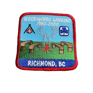 May include: Embroidered patch with a red border, featuring text "WOODWARDS LANDING 1980-2000" and "RICHMOND, BC". The patch depicts a campfire, wooden benches, and other camping-themed graphics on a blue and green background.