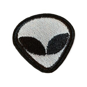 Pode incluir: Patch bordado com o rosto de um alienígena com grandes olhos pretos e uma cabeça cinza. O patch tem uma borda preta e foi projetado para ser passado a ferro ou costurado em roupas ou acessórios. O design é simples e gráfico.
