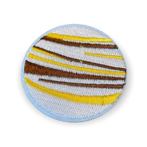 May include: Embroidered patch with a circular design. The patch features a light gray background with brown and yellow horizontal stripes. The patch has a light blue border.