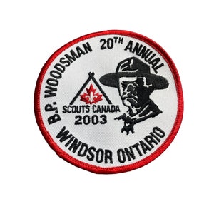 May include: A round patch with a white background and a red border. The patch features text that reads "B.P. WOODSMAN 20TH ANNUAL" and "WINDSOR ONTARIO". A black and white illustration of a man in a hat is also present.
