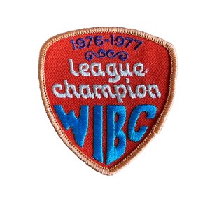 May include: A shield-shaped embroidered patch with a red background and a gold border. The patch features the text "1976-1977 League champion WIBG" in white and blue thread. The patch is likely a vintage sports or team emblem.