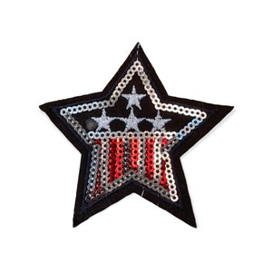 Puede incluir: Un parche negro en forma de estrella con un diseño patriótico. El parche presenta lentejuelas plateadas que delinean la estrella y rodean las estrellas interiores. Lentejuelas rojas y plateadas forman rayas y se ven tres estrellas plateadas.