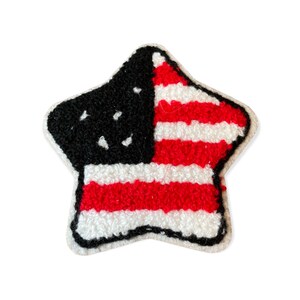 May include: A star-shaped patch with a design resembling the American flag. The patch features a black field with white stars in the upper left corner, and red and white stripes on the right. The edges are outlined in a cream color.