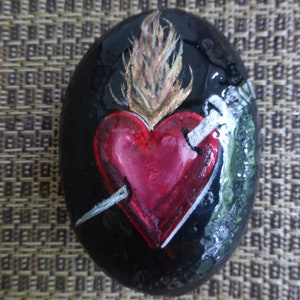 May include: A painted rock with a red heart pierced by a sword, a golden flame, and green foliage. The heart is painted in a realistic style with a three-dimensional effect.