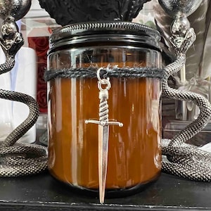 Peut inclure: Un bocal en verre brun contenant une bougie, avec une corde noire nouée autour du haut. Un pendentif en forme de dague argentée est suspendu à la corde. Le bocal à bougie est posé sur une surface noire.