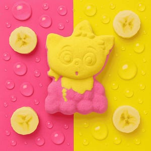 May include: A yellow and pink cat-shaped bath bomb with a surprised expression, set against a split background of pink and yellow. Sliced bananas and water droplets are scattered around the bath bomb, creating a playful and colourful composition.