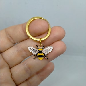 May include: A gold-coloured keychain with a bee charm. The bee is yellow and black with white wings.