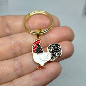 May include: Gold key ring with a white rooster charm. The rooster has a red comb and wattle, black feathers on its head and tail, and white feathers on its body.