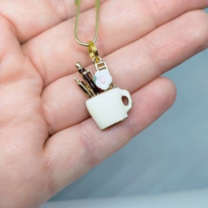 May include: Gold charm featuring a white ceramic mug with a gold handle, filled with gold and brown pencils and a pink flower.