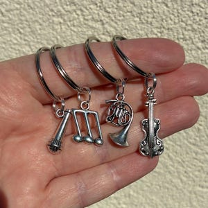 May include: Silver-toned keychains with musical instrument charms. The charms include a microphone, musical notes, a French horn, and a violin. The keychains are held in a hand against a neutral background.