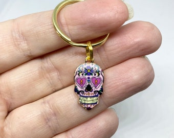 May include: A gold key chain with a pink and purple sugar skull charm. The skull has pink hearts for eyes and a floral design.
