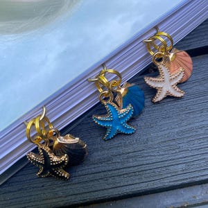 May include: Three gold-coloured metal stitch markers with seashell and starfish charms. The starfish are in black, white, and blue with gold accents. The charms are attached to gold rings, ideal for knitting or crochet projects.