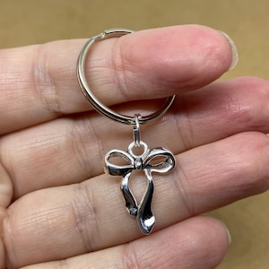 May include: A silver-coloured keyring featuring a bow pendant. The bow has a detailed design with flowing ribbon ends. The keyring ring is attached to the bow pendant by a small loop.
