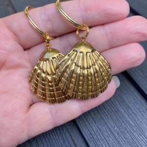 May include: A pair of gold-coloured seashell earrings. Each earring has a detailed, textured shell pendant suspended from a circular hoop. The earrings are designed to be worn, adding a touch of coastal elegance.