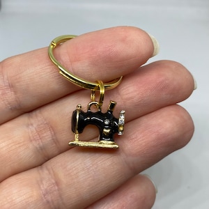 May include: Gold key chain with a black and gold sewing machine charm. The sewing machine has a gold base and a black body with a gold accent. The sewing machine has a row of clear rhinestones around the top.
