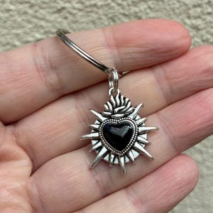 May include: Silver-toned keychain with a pendant featuring a black heart at the centre, encircled by a starburst design. The keychain is held in a hand, highlighting the pendant's intricate details. The design is reminiscent of religious iconography.