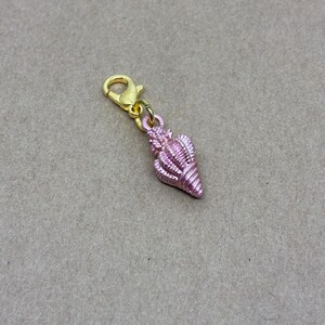 May include: A pink seashell charm with a gold-coloured clasp. The seashell is detailed with ridges and a textured surface. The charm is designed to be attached to a bracelet or necklace. The background is a neutral tan colour.