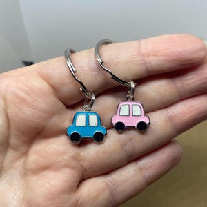 May include: Two car-shaped keychains. One is blue with a white window frame, the other is pink with a white window frame. Both have black wheels and silver key rings. The keychains are small and colorful.
