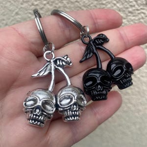 May include: Two keyrings, each featuring a pair of skull-shaped cherries. One is silver, the other black. The skulls have detailed features and are attached to a stem and leaf design. The keyrings are held in a hand.