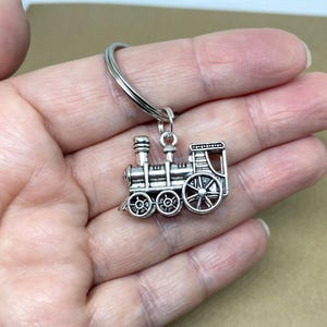 May include: A silver-coloured train engine keyring. The miniature train has detailed wheels, smokestacks, and a cab. The keyring is a small, decorative accessory.