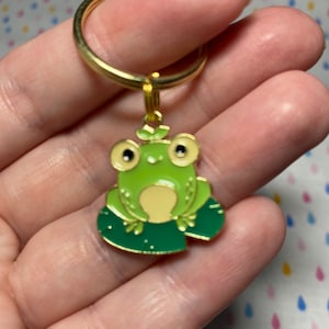 May include: A gold-coloured keychain featuring a green frog sitting on a green lily pad. The frog has large, black eyes and a small, green leaf on its head.