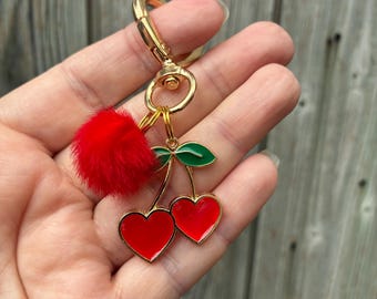 May include: A gold-toned metal keychain featuring two red heart-shaped charms attached to a green enamel leaf accent. The keychain includes a small, fluffy red pom-pom and a gold lobster clasp for attachment, creating a decorative accessory.